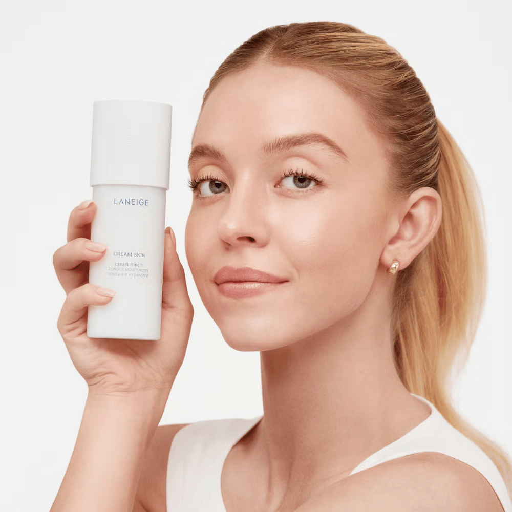 Henkilö nostaa Laneige Cream Skin Cerapeptide Refiner valkoisessa pullossa. Henkilö hymyilee ja esittelee tuotteen, jota markkinoidaan kosteuttavana kasvovetenä ja ihonhoitotuotteena.