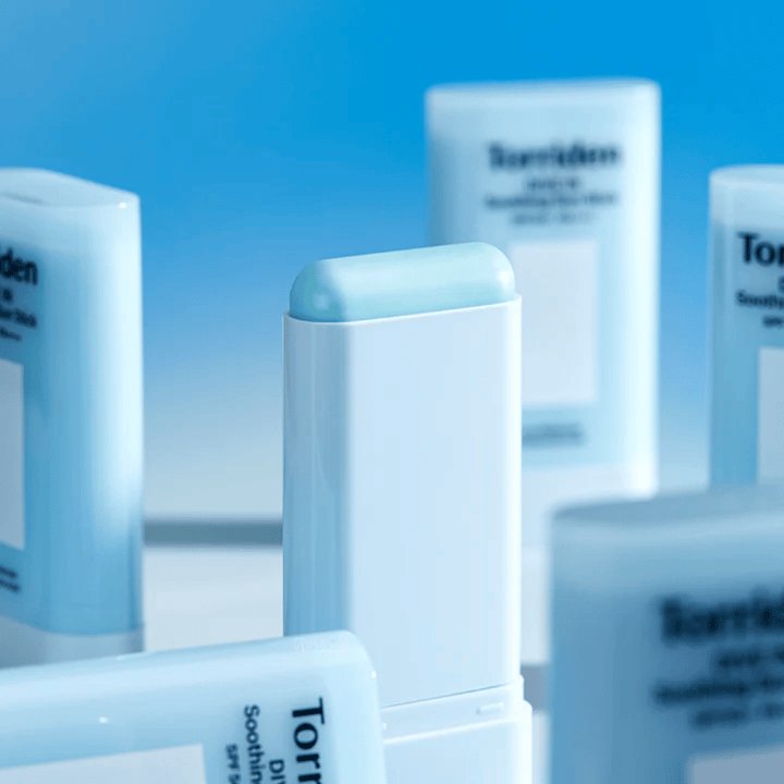 Lähikuva tuotteesta Torriden DIVE IN Soothing Sun Stick, joka näyttää pehmeän aurinkosuojakoostumuksen stick-muodossa useiden tuotteiden kanssa taustalla.