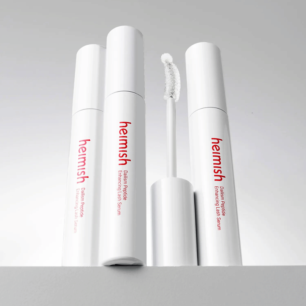 Useita tuotteiden Heimish Dailism Peptide Enhancing Lash Serum pakkauksia esitellään yhdessä kaarevan applikaattoriharjan kanssa. Kuva korostaa tuotteen eleganttia muotoilua ja tarkkuusharjaa helppoa levitystä varten.