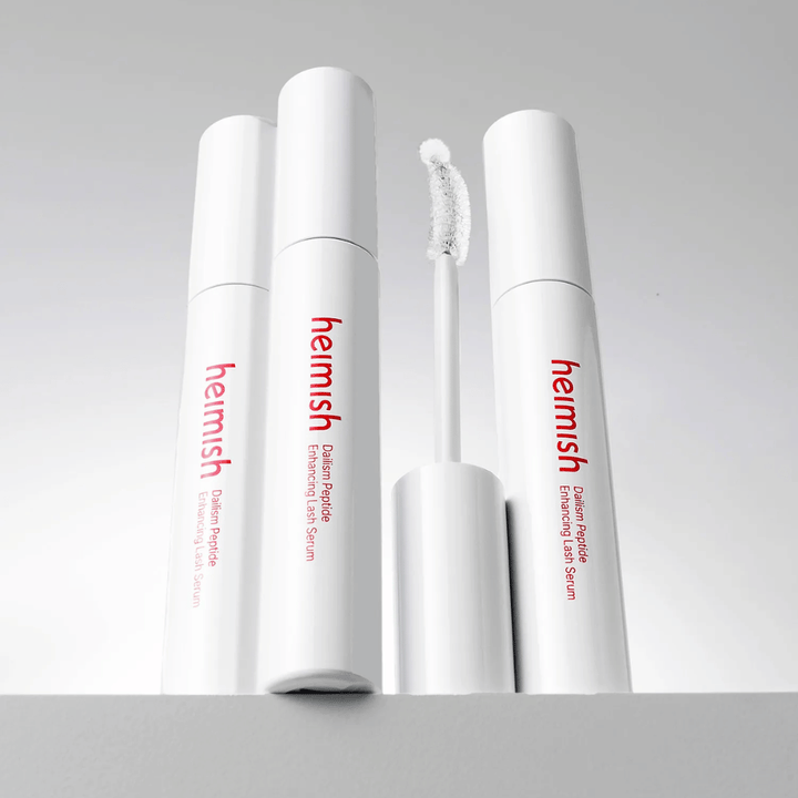 Useita tuotteiden Heimish Dailism Peptide Enhancing Lash Serum pakkauksia esitellään yhdessä kaarevan applikaattoriharjan kanssa. Kuva korostaa tuotteen eleganttia muotoilua ja tarkkuusharjaa helppoa levitystä varten.