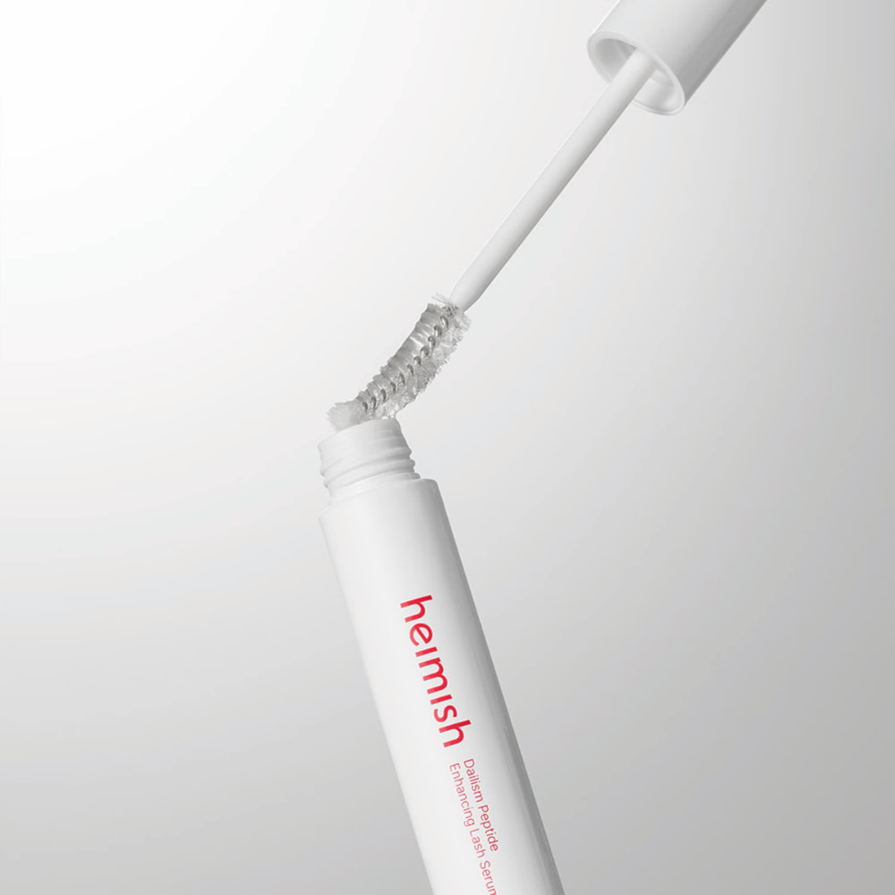 Lähikuva tuotteiden Heimish Dailism Peptide Enhancing Lash Serum applikaattorista. Harja vedetään ylös valkoisesta säiliöstä, ja se näyttää läpinäkyvän seerumikoostumuksen, joka levitetään ripsille.