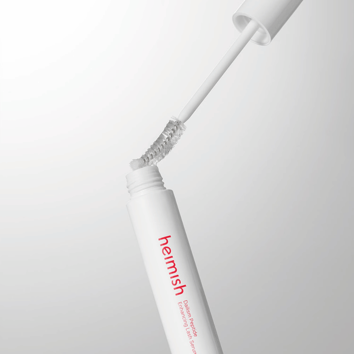 Lähikuva tuotteiden Heimish Dailism Peptide Enhancing Lash Serum applikaattorista. Harja vedetään ylös valkoisesta säiliöstä, ja se näyttää läpinäkyvän seerumikoostumuksen, joka levitetään ripsille.