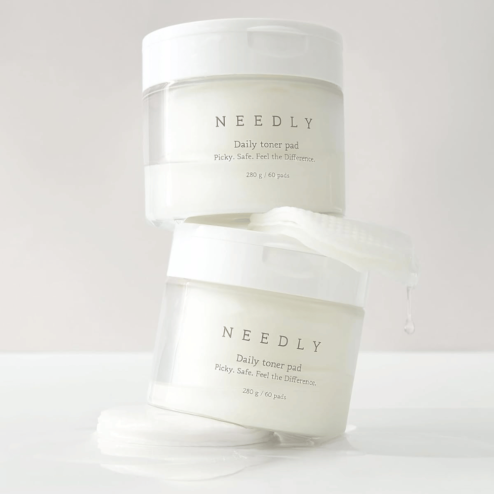 Kaksi purkkia, joissa on Needly Daily Toner Pad pinottuna toistensa päälle. Purkit ovat valkoisia minimalistisella tekstillä ja sisältävät 60 kasvovettä kumpikin. Yksi kasvovesi on osittain ulkona ylemmästä purkista nesteen tippuessa siitä.