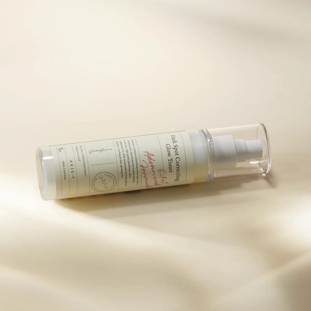 Makaava pullo "AXIS-Y Dark Spot Correcting Glow Toner" asetettuna vaalealle pinnalle. Pullo on läpinäkyvä, siinä on valkoinen etiketti ja valkoinen suihkepumppu. Etiketti näyttää tietoa tuotteen kirkastavista ja kosteuttavista ominaisuuksista sekä sisältää avainainesosia kuten glutationi ja niasiiniamidi.
