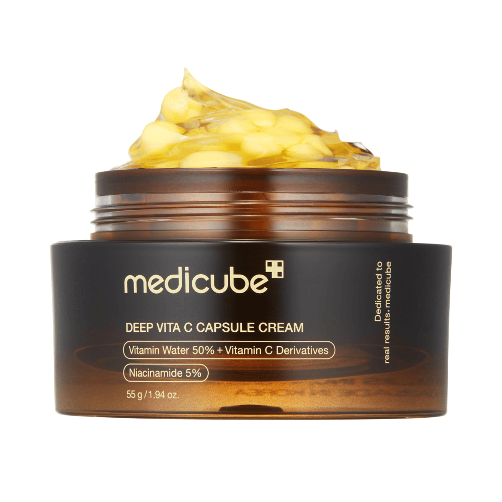 Lähikuva "Medicube Deep Vita C Capsule Cream," joka näyttää voiteen koostumuksen ja C-vitamiinikapselit. Pakkaus on tummanruskea kultatekstillä ja kuvailee C-vitamiinivettä (50 %) ja niasiiniamidia (5 %), mikä antaa visuaalisen esityksen tuotteen kosteuttavista ja ihoa parantavista ominaisuuksista.