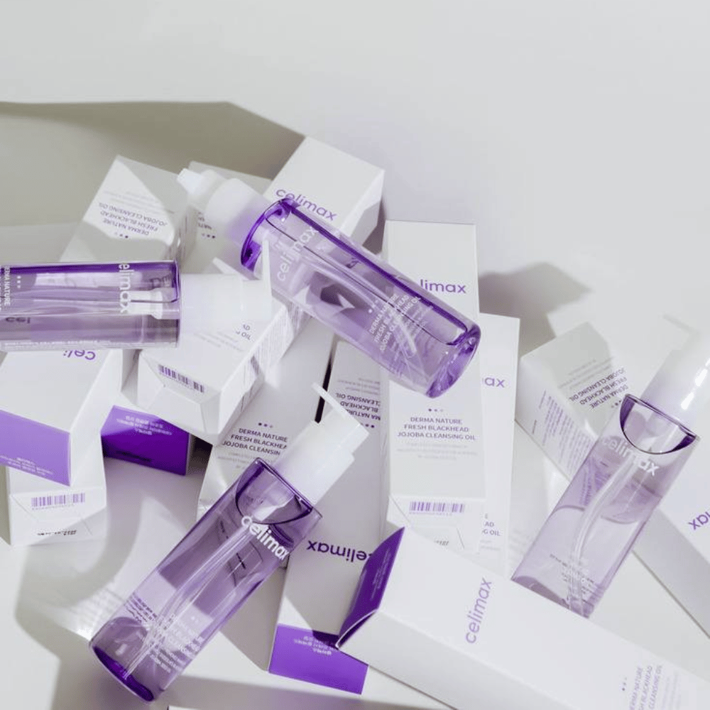 Celimax Derma Nature Fresh Blackhead Jojoba Cleansing Oil violetissa pulloissa sijoitettuna valkoisten tuotepakkausten joukkoon, joissa on violetit yksityiskohdat.