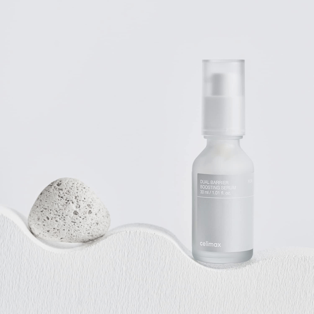 Celimax Dual Barrier Boosting Serum asetettu kankaalle kiven kanssa, rauhallinen tuotekuva, joka viestii kosteutuksesta ja vahvistetusta ihon suojamuurista.
