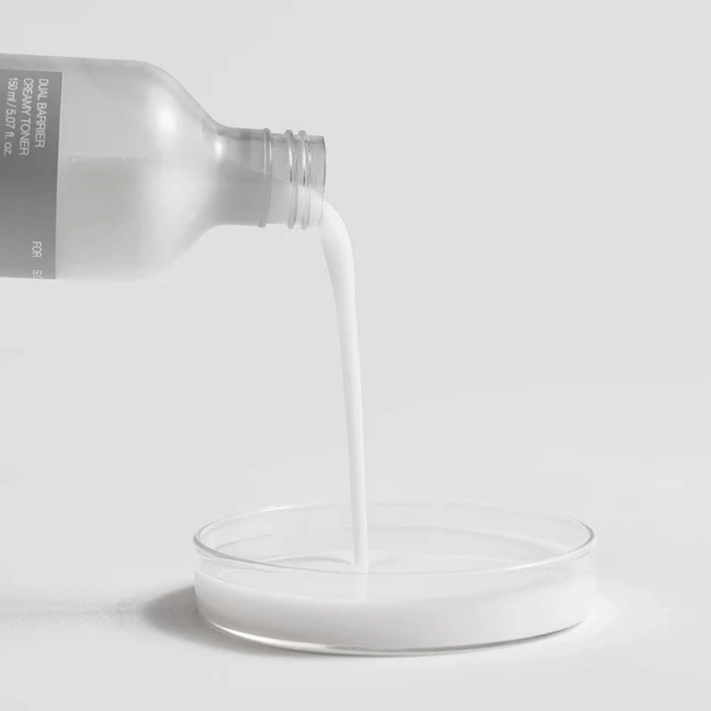 Celimax Dual Barrier Creamy Toner kaadetaan varovasti pullosta pyöreään laboratoriokaivoon, sen maitomainen ja ravitseva koostumus selvästi keskiössä.