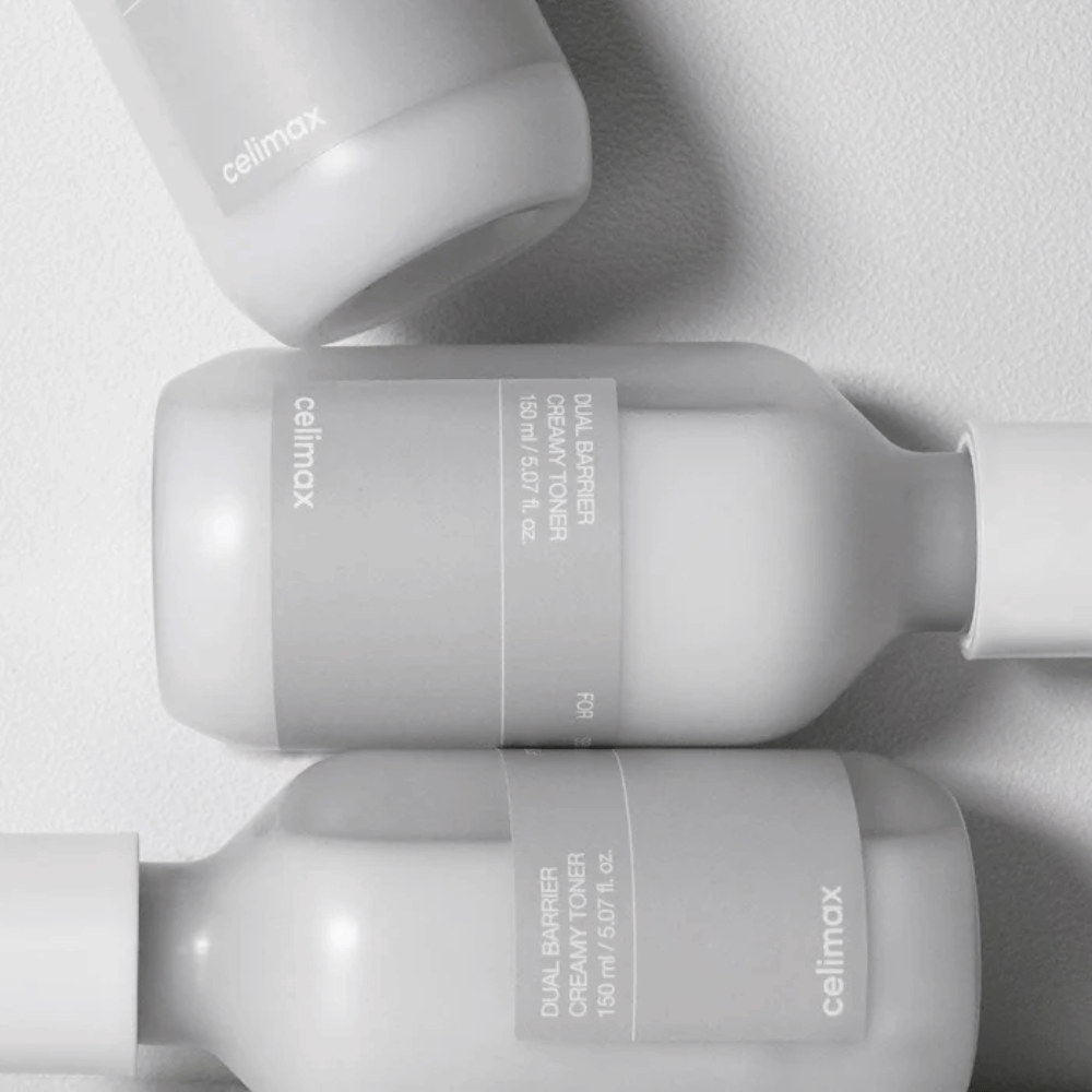Kolme makuullaan olevaa pulloa Celimax Dual Barrier Creamy Toner minimalistisessa muotoilussa harmaalla etiketillä ja pehmeällä valolla – täydellinen herkälle ja kuivalle iholle.