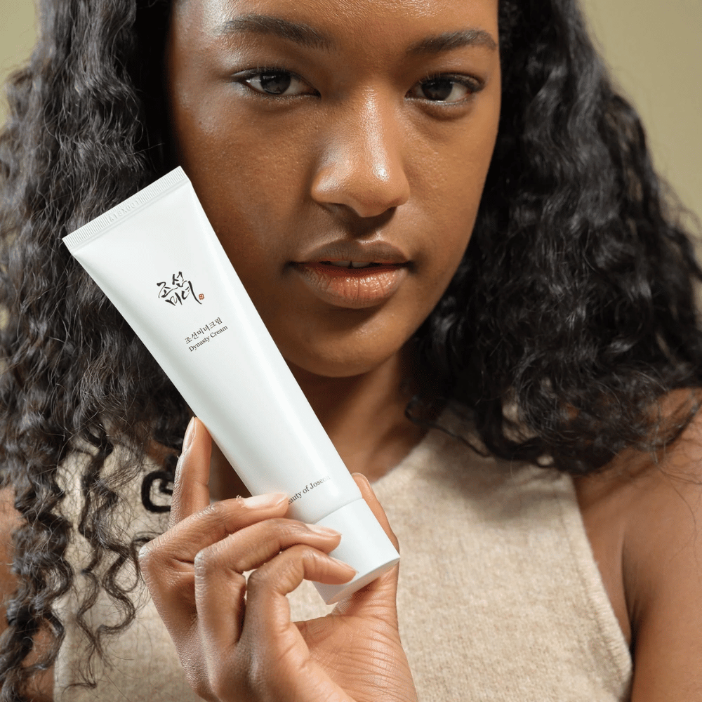 Kiharatukkainen nainen pitää "Beauty of Joseon Dynasty Cream" -tuubia lähellä kasvojaan, mikä korostaa tuotetta. Voide, joka on esitelty tyylikkäässä valkoisessa tuubissa, on suunniteltu ravitsemaan ja nuorentamaan ihoa, korostaen sen houkuttelevuutta eri ihotyypeille.