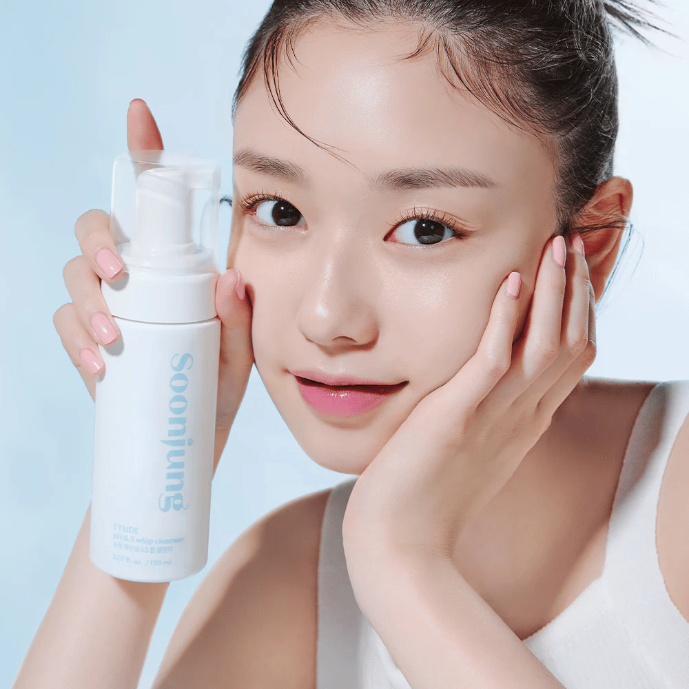 Nuori nainen, jolla on kirkas iho, pitää valkoista pumppupulloa, jossa on teksti "Soon Jung pH 6.5 Whip Cleanser" ETUDE. Hän hymyilee kameralle kevyellä meikillä ja raikkaalla ilmeellä vaaleansinisellä taustalla.