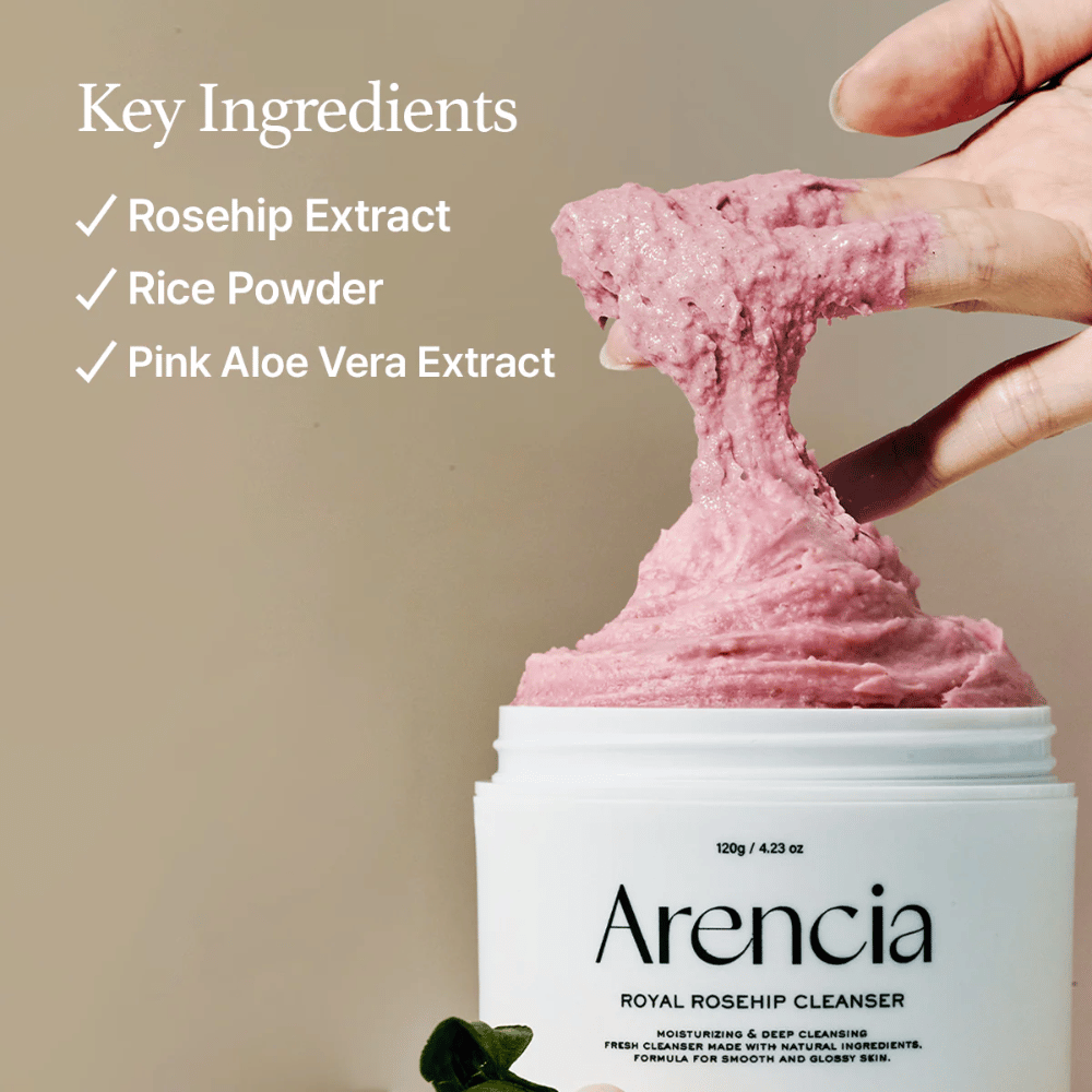 Käsi nostaa vaaleanpunaista, pöyheää puhdistuskoostumusta purkista Arencia Royal Rosehip Cleanser, vasemmalla ainesosaluettelo: ruusunmarjauute, riisijauhe ja vaaleanpunainen aloe vera -uute.