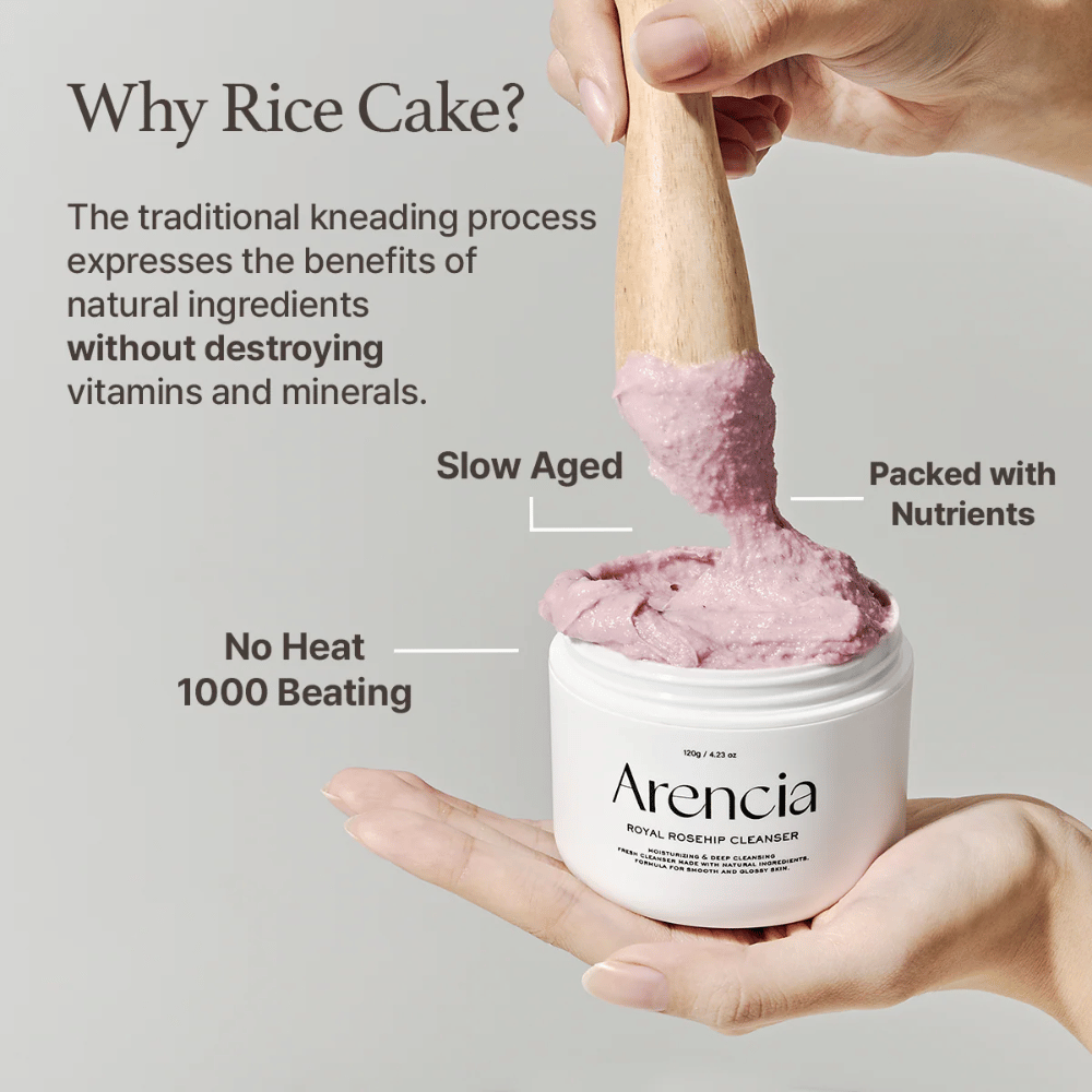 Käsi pitelee avointa purkkia Arencia Royal Rosehip Cleanser, jossa on vaaleanpunainen, mochia muistuttava puhdistuskoostumus, joka nostetaan puulastalla – näyttää tuotteen koostumuksen ja perinteisen valmistusmenetelmän.