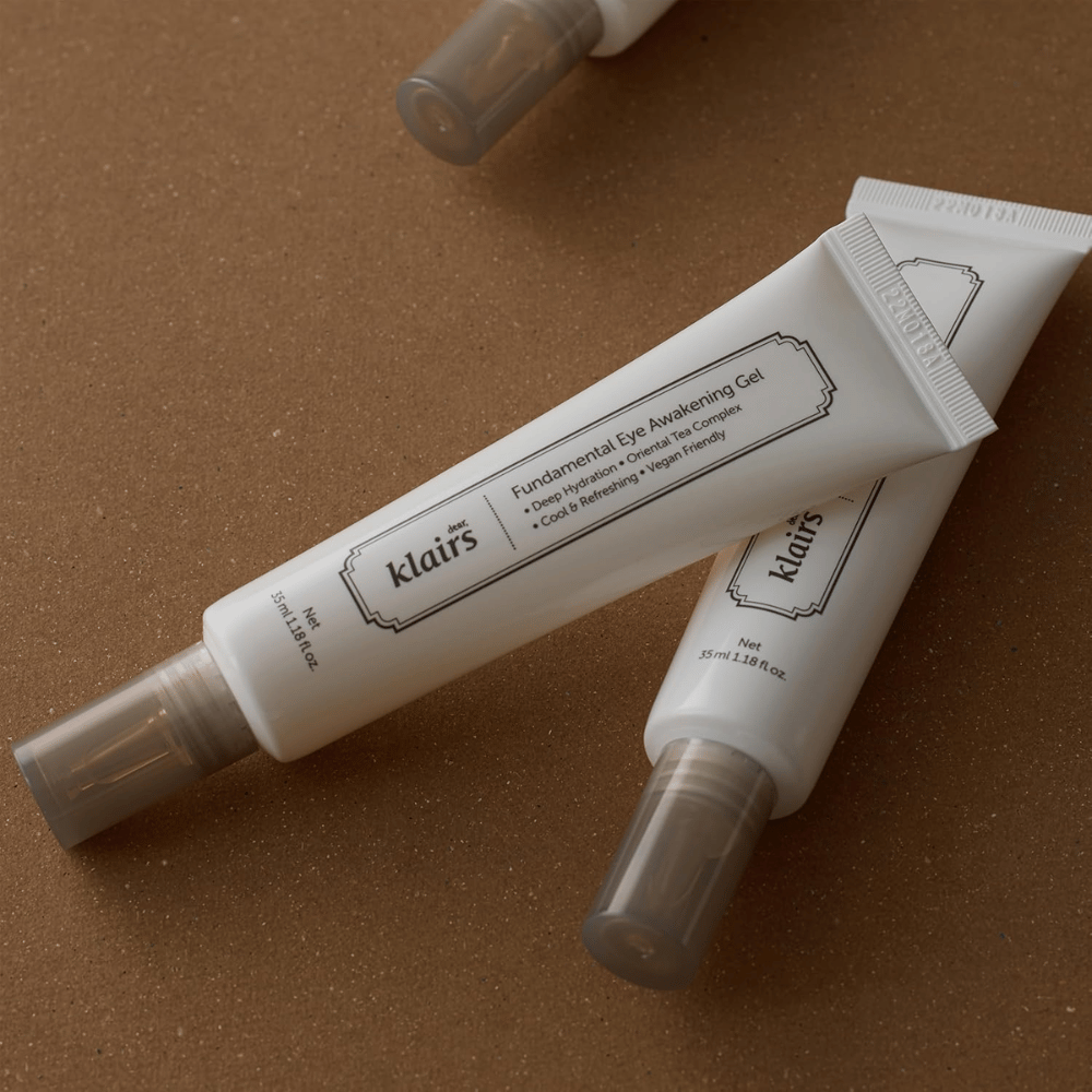 Lifestyle-kuva, jossa kaksi tuubia tuotetta Klairs Fundamental Eye Awakening Gel on asetettu lämpimän ruskeaa taustaa vasten. Sommittelu korostaa tuotteen eleganttia muotoilua ja premium-tuntua samalla, kun huomio kohdistuu silmänympärysalueen hoitoon ja kosteutukseen.