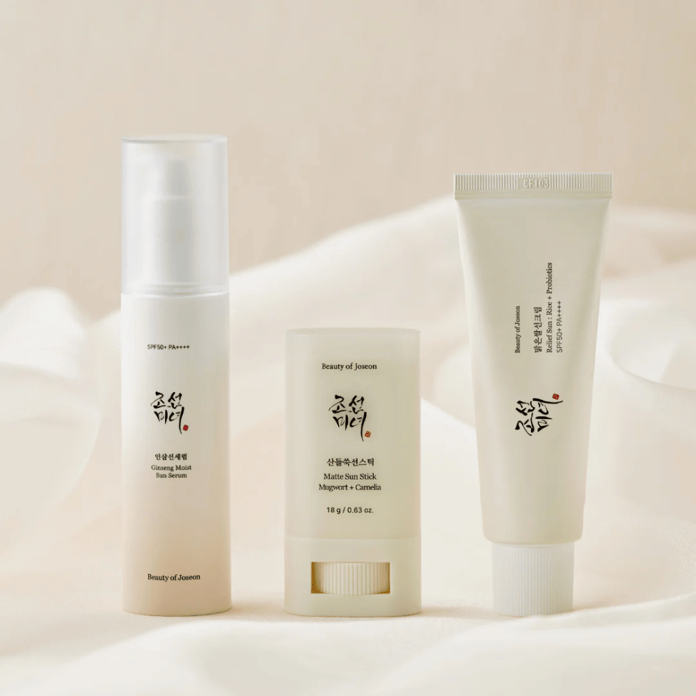 Kolme aurinkosuojatuotetta Beauty of Joseon – Ginseng Moist Sun Serum, Matte Sun Stick ja Relief Sun Rice + Probiotics – ovat rivissä kermanvärisellä kankaalla pehmeässä valossa.