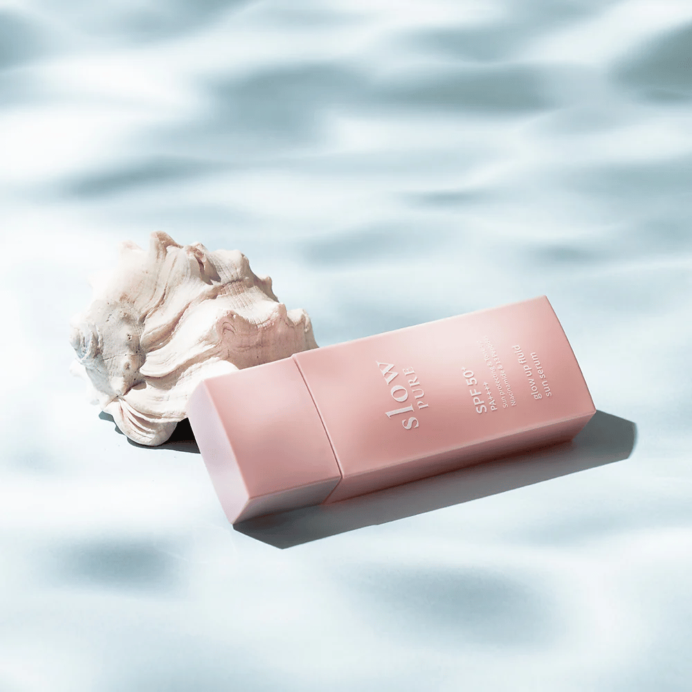 Slow Pure Glow Up Fluid Sun Serum simpukankuorien ja vesiefektin vieressä, korostaa aurinkosuojaa ja hehkua.