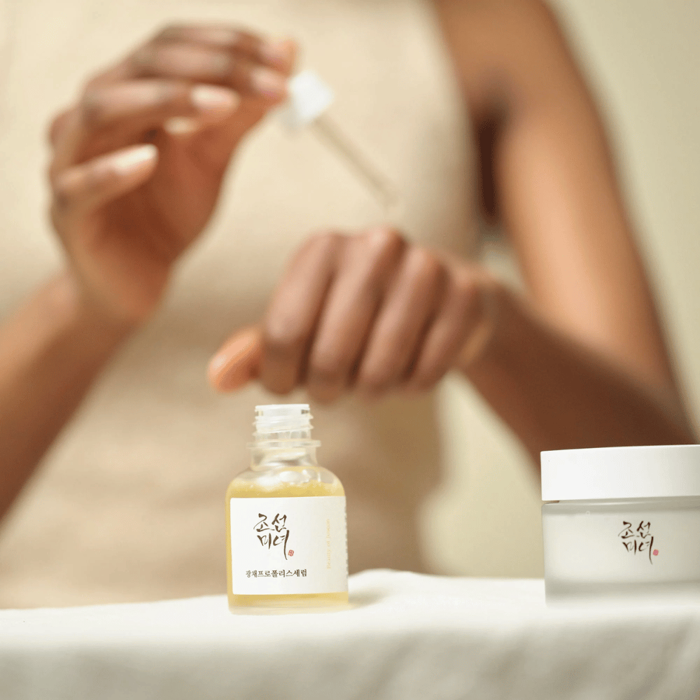 Henkilö, joka levittää Beauty of Joseon Glow Serum pipetillä lasipullosta, viereisellä pöydällä saman sarjan purkki.