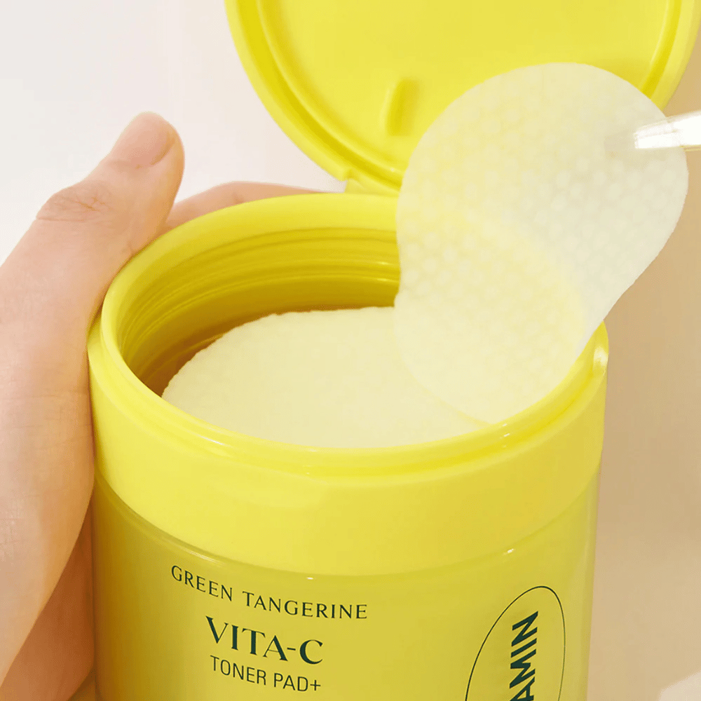 Käsi käyttää muovipinsettejä nostaakseen tekstuurillisen kasvovesilapun vaaleankeltaisesta säiliöstä, jossa on merkintä "GREEN TANGERINE VITA-C TONER PAD+" merkiltä "gooda".
