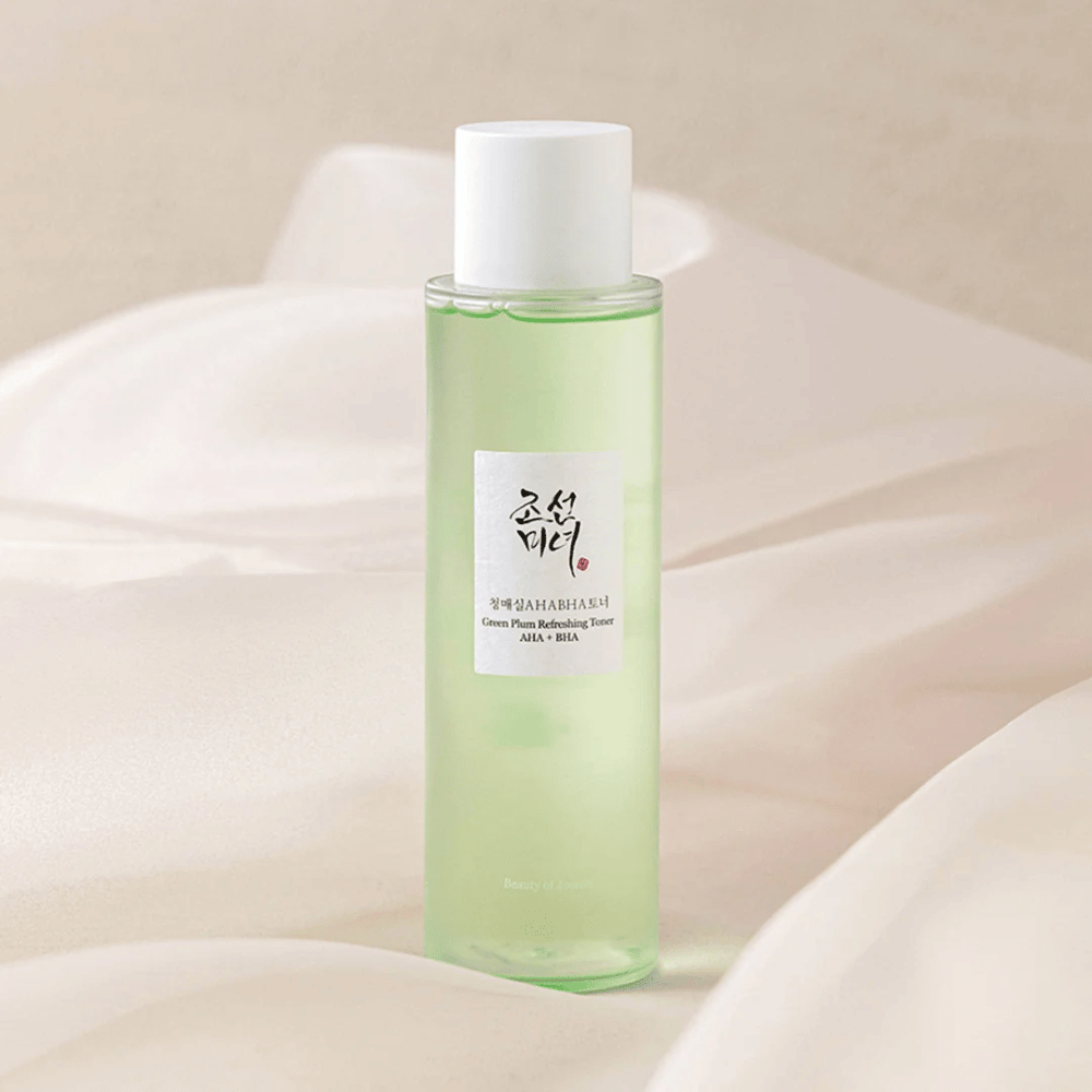 "Green Plum Refreshing Toner" AHA + BHA -pullo seisoo pehmeällä, vaalealla taustalla. Tuotteessa on vihreä neste, joka heijastaa raikkautta ja luonnollisuutta. Etiketti on valkoinen mustilla ja punaisilla yksityiskohdilla, mikä antaa tyylikkään ja puhtaan ilmeen.