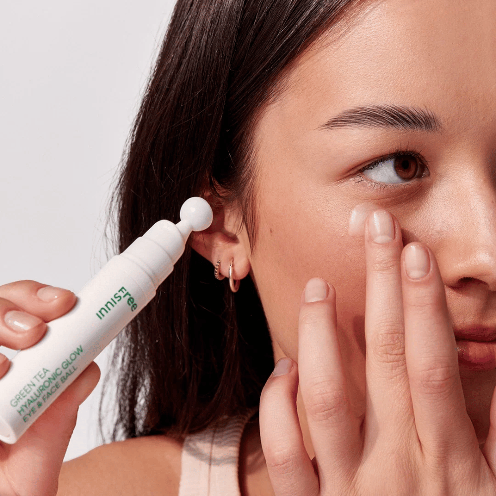 Henkilö levittää Innisfree Green Tea Hyaluronic Glow Eye & Face Ball iholleen silmien alle. Tuotteessa on valkoinen, pyöreä levityspää ja se on valkoisessa pullossa, jossa on vihreä teksti. Käyttäjän kasvot näkyvät osittain ja keskittyvät tuotteen levitykseen.