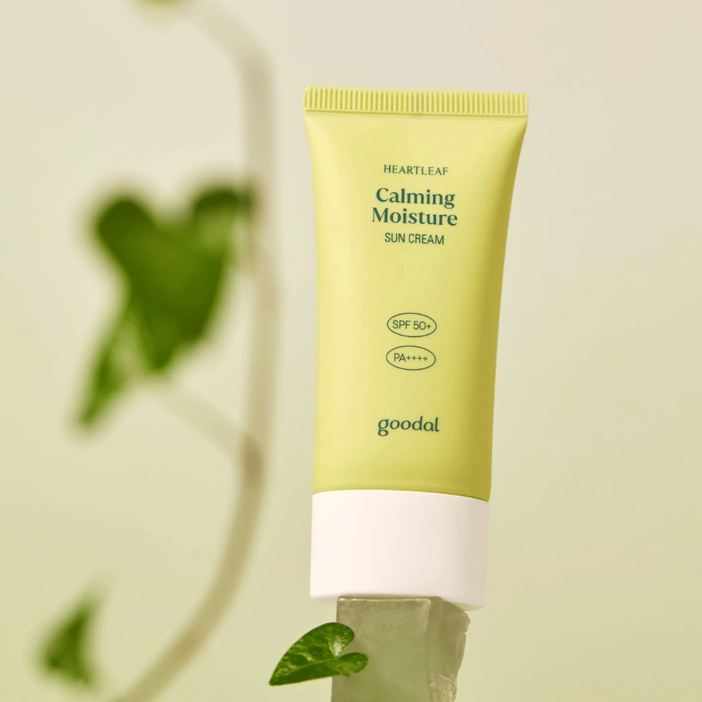 Goodal Heartleaf Calming Moisture Sun Cream SPF50+ PA++++ sijoitettuna lasilevylle, taustalla vihreä kasvi – rauhoittava aurinkovoide herkälle iholle.