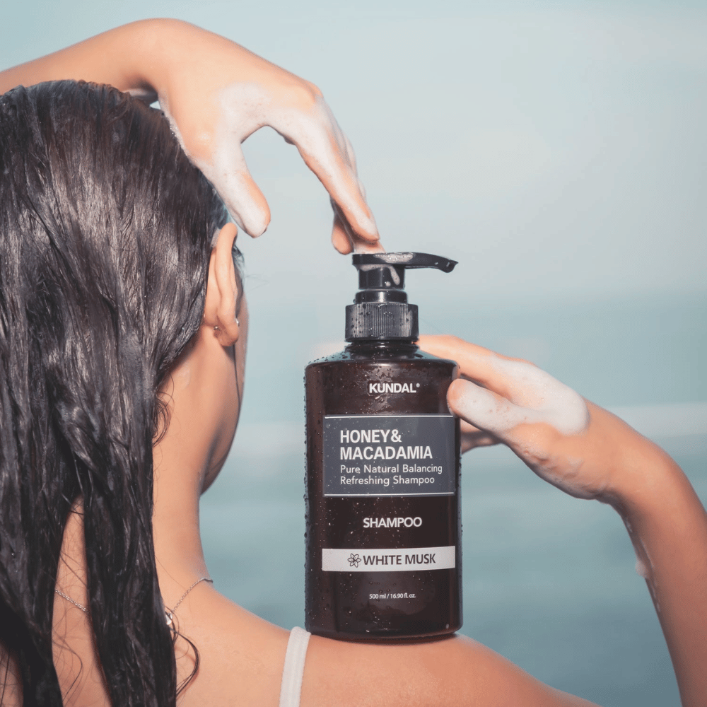 Nainen, jolla on märät hiukset, levittää vaahtoa ja pitää KUNDAL Honey & Macadamia Nature Shampoo White Musk merta vasten.