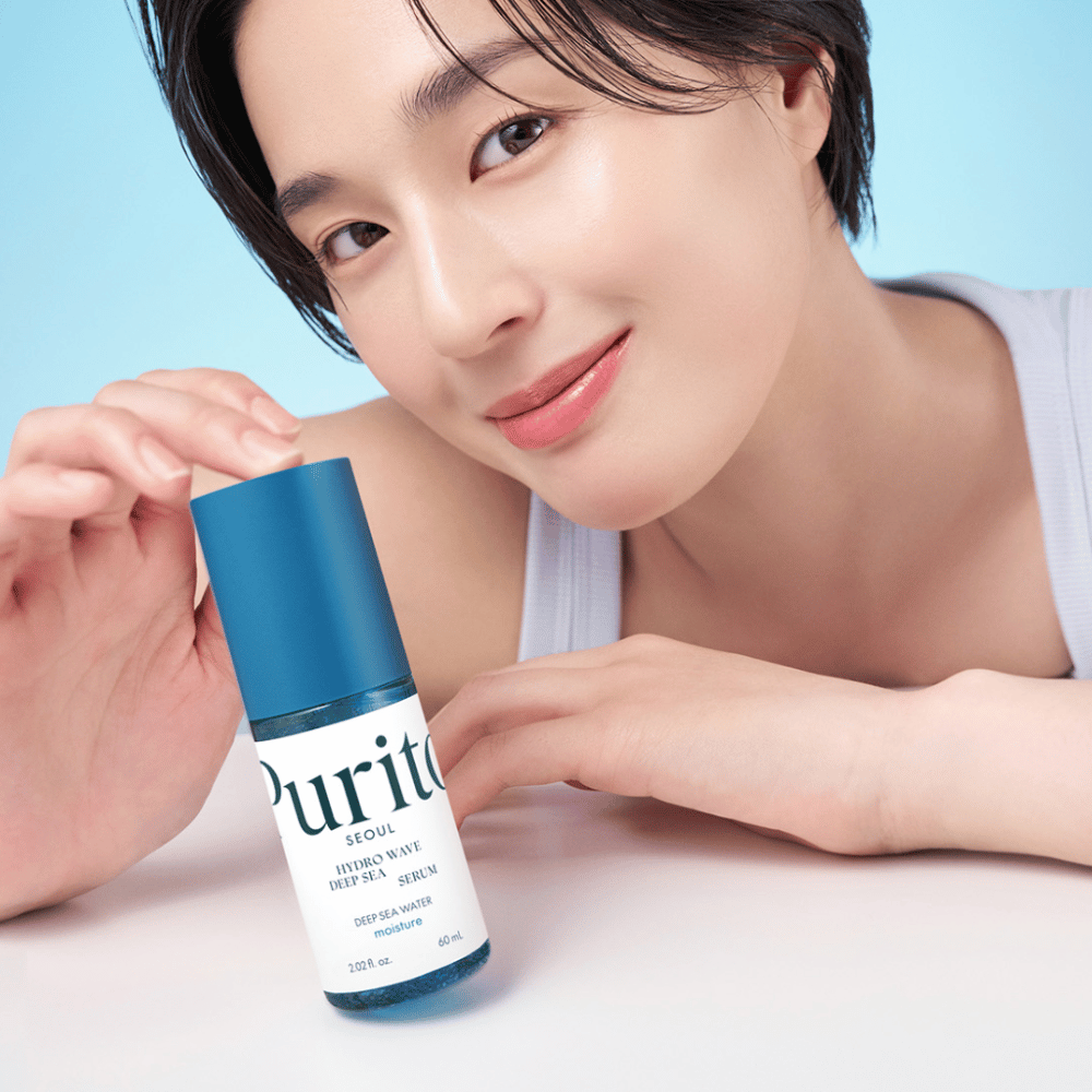 Nainen, jolla on lyhyet hiukset, pitää käsissään sinistä seerumia Purito, Hydro Wave Deep Sea Serum. Hänen ihonsa näyttää säteilevältä ja kosteutetulta, mikä korostaa tuotetta keskipisteessä. Tausta on vaaleansininen, mikä luo raikkaan ja puhtaan tunteen. Tuote keskittyy ihonhoitoon.