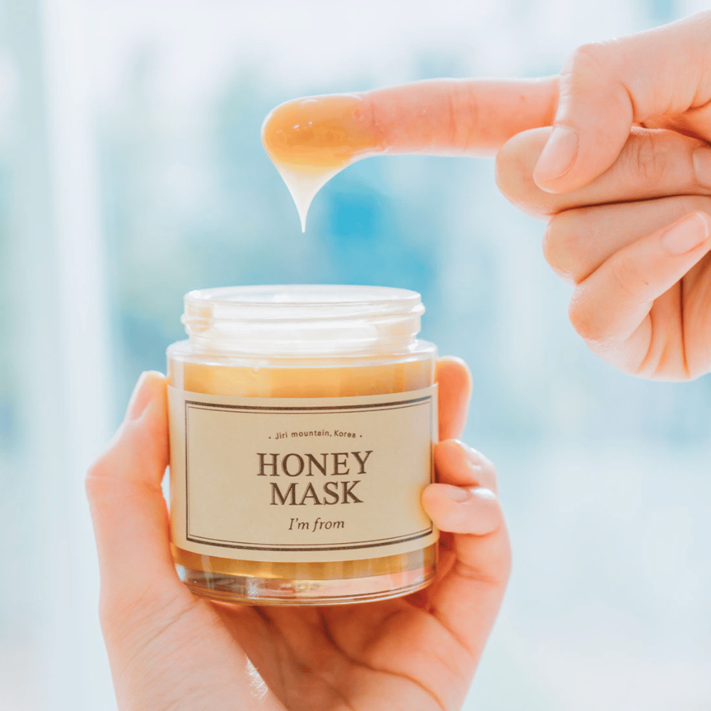 Käsi pitää avointa purkkia "I'm from Honey Mask", ja sormi kastetaan kultaiseen naamioon vaaleaa taustaa vasten.