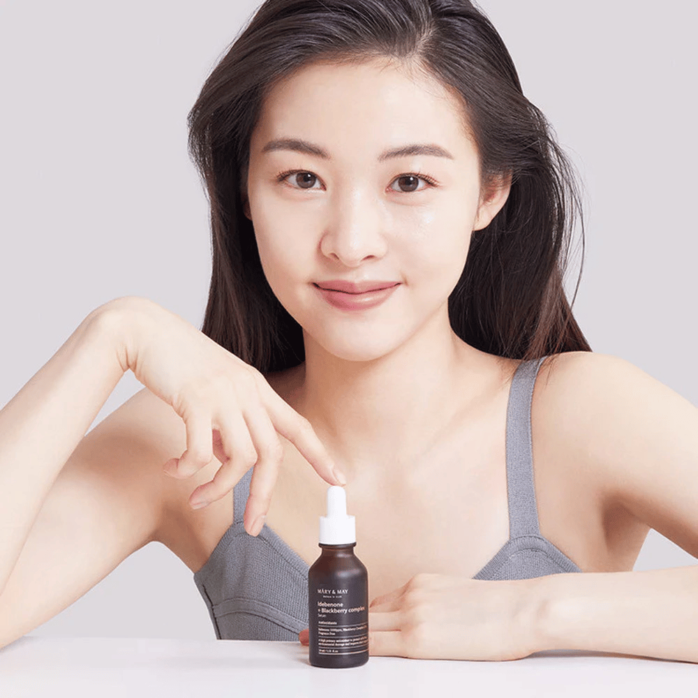 Pitkät, tummat hiukset omaava nainen pitää kädessään MARY & MAY Idebenone + Blackberry Complex Serum, tumma lasipullo pipetillä. Hänellä on iloinen hymy ja hän osoittaa pullon suuntaan toisella kädellään. Nainen on pukeutunut harmaaseen, hihattomaan toppiin ja näyttää olevan valoisassa ja yksinkertaisessa ympäristössä, mikä korostaa tuotteen keskittymistä ihonhoitoon ja kauneuteen.