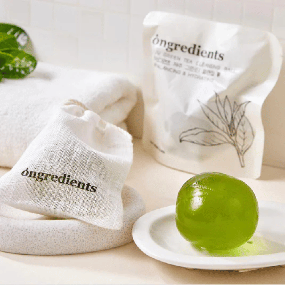 Óngredients Jeju Green Tea Cleansing Ball asetettuna lautaselle pakkauksen ja pyyhkeen kanssa – hellävarainen puhdistus Jejun vihreällä teellä.