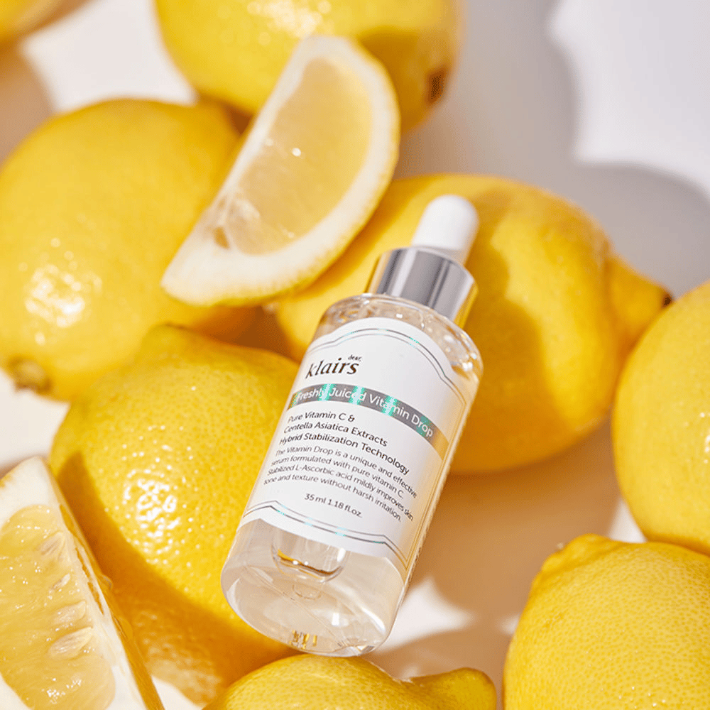 Klairs Freshly Juiced Vitamin Drop seerumipullo, jota ympäröivät tuoreet sitruunanpuolikkaat ja sitruunaviipaleet vaalealla pinnalla, korostaa ainesosia.
