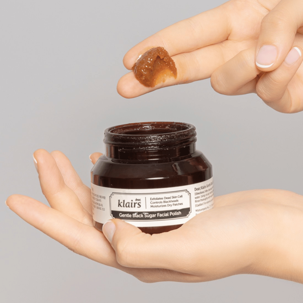 Kädet pitävät avointa purkkia Klairs Gentle Black Sugar Facial Polish ja näyttävät tuotteen sormella, harmaata taustaa vasten.