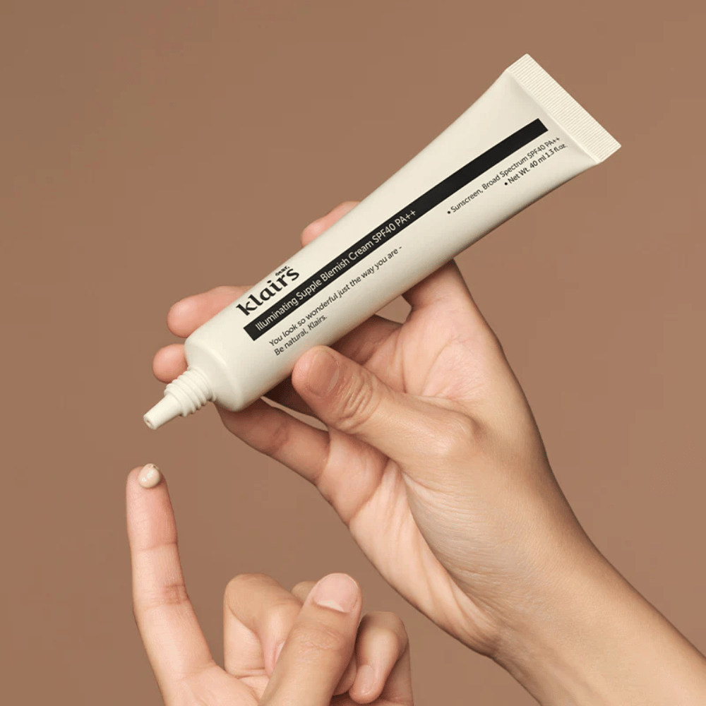Käsi pitää putkiloa Klairs Illuminating Supple Blemish Cream SPF40 PA++ ja puristaa pienen määrän voidetta sormelle, ruskeaa taustaa vasten.