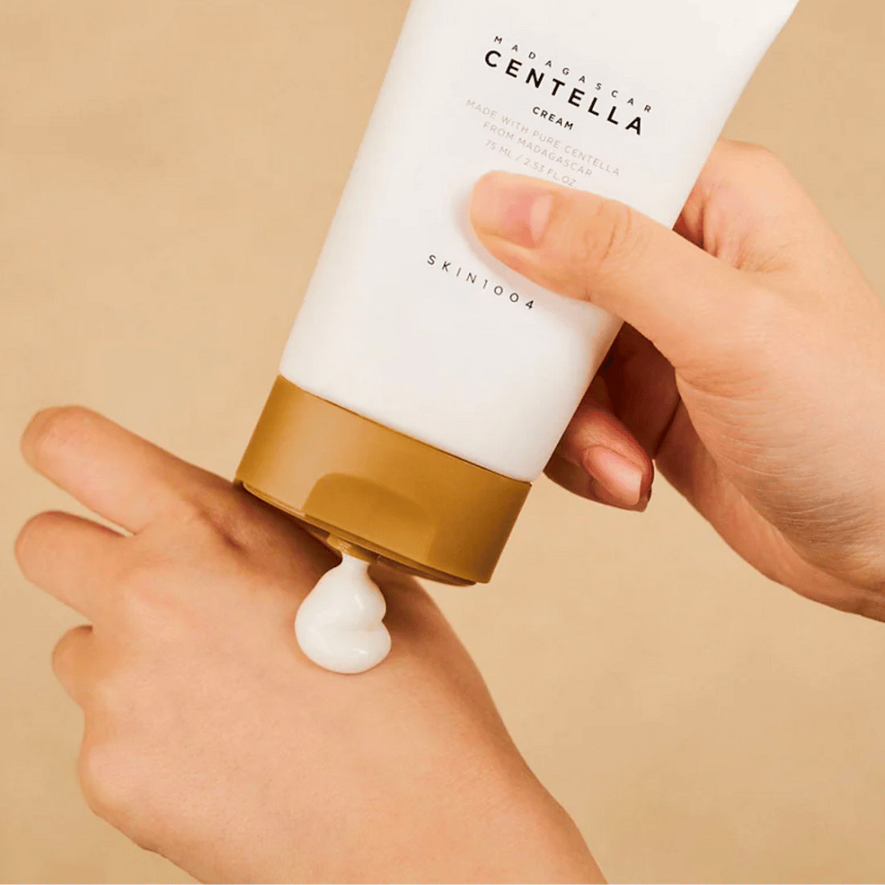 Kuva näyttää henkilön, joka pitelee "Madagascar Centella Cream" tuubia SKIN1004. Se on ihovoide, joka on valmistettu puhtaasta Madagaskarin Centellasta, 75 ml / 2,53 fl.oz. koossa. Voide levitetään käteen ja on tarkoitettu tarjoamaan ravintoa ja kosteutusta iholle.