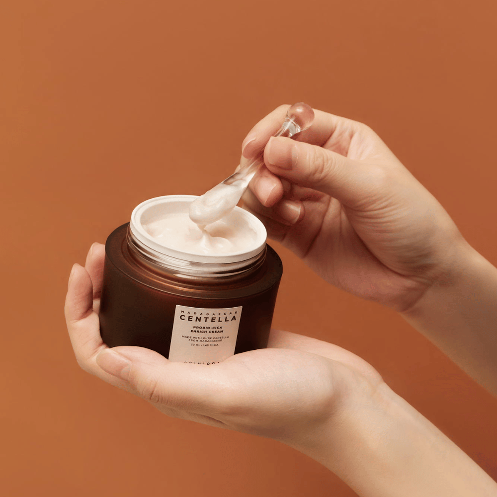 Kädet pitävät avointa purkkia Madagascar Centella Probio-Cica Enrich Cream ja lastaa voiteella yhtenäistä terrakotanväristä taustaa vasten.