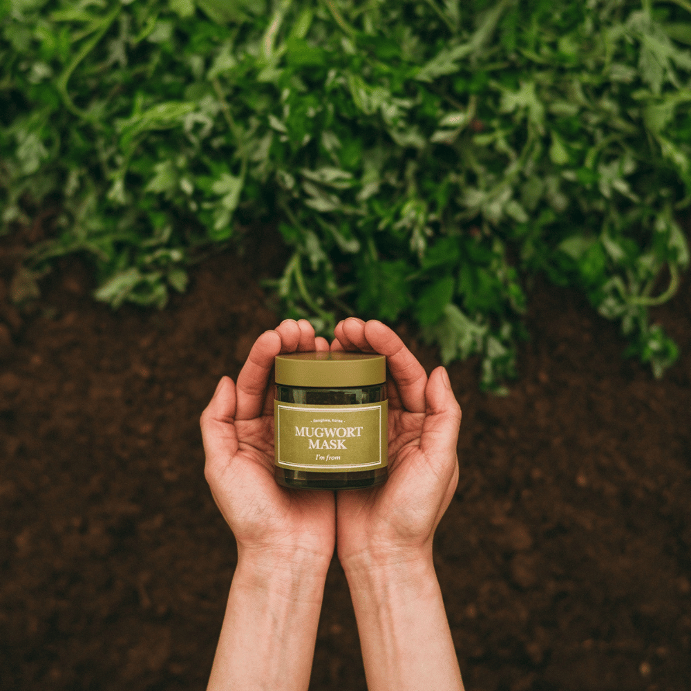 Kädet pitävät I'm From Mugwort Mask taustalla maata ja tuoreita vihreitä yrttejä, korostaen tuotteen luonnollisia ainesosia.
