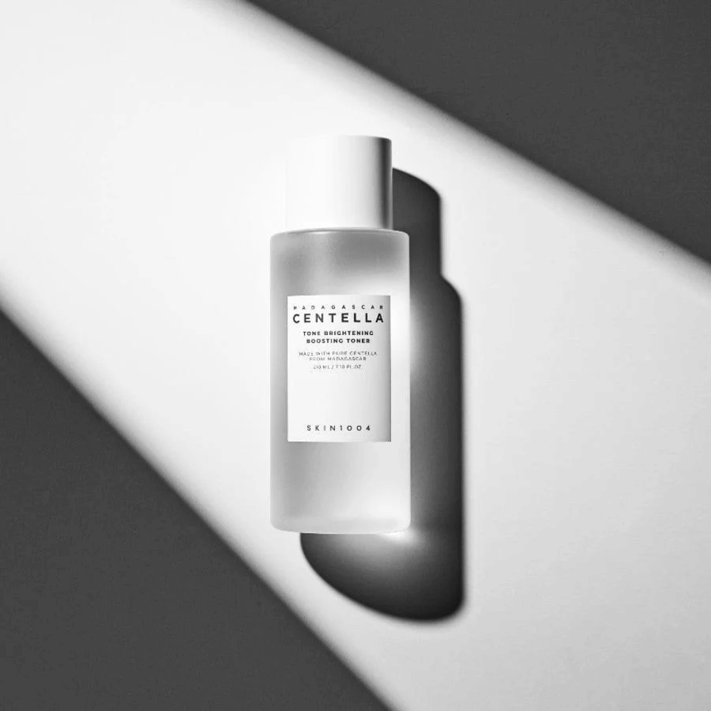 Pullo SKIN1004 Madagascar Centella Tone Brightening Boosting Toner makaamassa valkoisella pinnalla dramaattisen varjon kanssa mustavalkoisessa valossa.