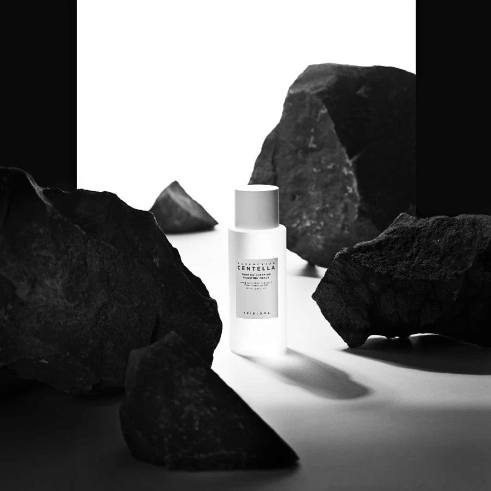 Valkoinen pullo SKIN1004 Madagascar Centella Tone Brightening Boosting Toner sijoitettuna dramaattisesti suurten mustien kivien joukkoon valkoista taustaa vasten.