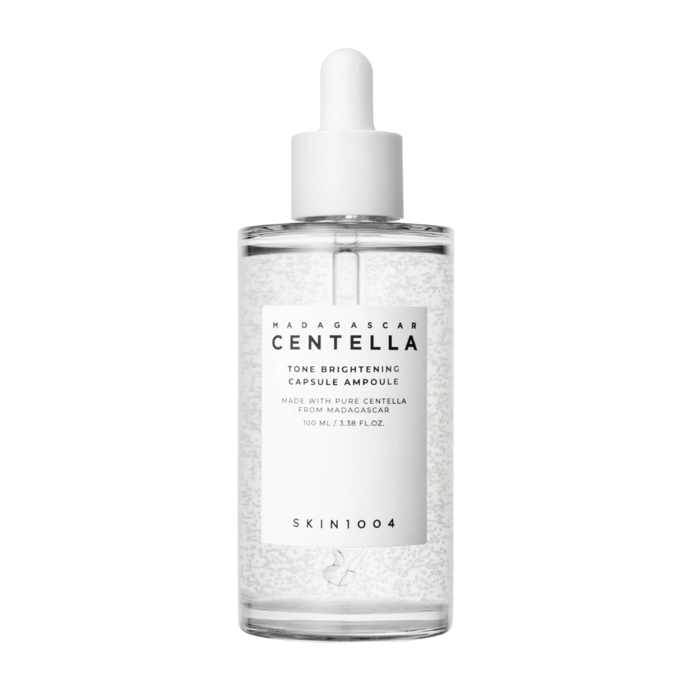 Kuva näyttää pullon "SKIN1004 Madagascar Centella Tone Brightening Capsule Ampoule". Se on läpinäkyvä pullo, jossa on valkoinen tippa-applikaattori. Etiketissä lukee, että tuote sisältää puhtaita centella-aineksia Madagaskarilta. Pullon sisältö näyttää sisältävän pieniä kapseleita tai partikkeleita, jotka voivat olla osa sen kirkastavia ihonhoito-ominaisuuksia. Pullo sisältää 100 ml / 3.38 fl.oz. tuotetta.