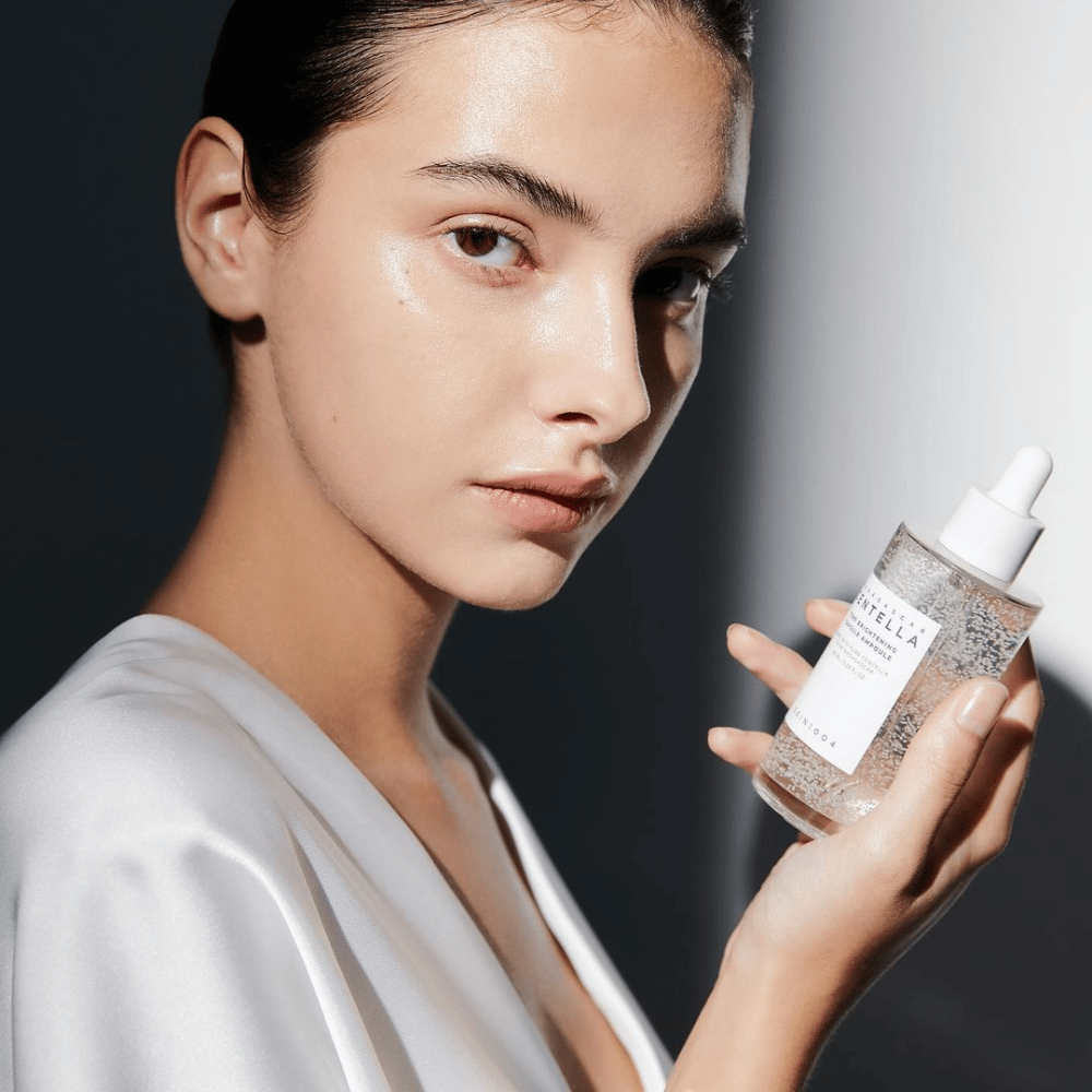 Kuvassa näkyy henkilö, joka pitää käsissään pulloa, jossa on tippa-applikaattori. Pullo on merkitty "SKIN1004 Madagascar Centella Tone Brightening Capsule Ampoule". Henkilöllä on hohtava ihonsävy, joka heijastaa valoa, mikä viittaa mahdolliseen tuotteen vaikutukseen.