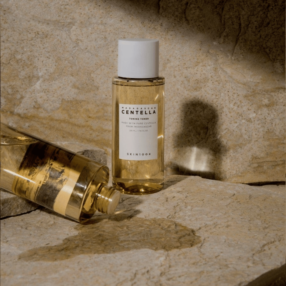 Kuva näyttää pullon "Madagascar Centella Toning Toner" SKIN1004. Se on läpinäkyvä pullo täynnä kellertävää nestettä, luultavasti ihonhoitotuotteen toner. Siinä lukee, että se on valmistettu puhtaasta Madagaskarin centellasta ja sisältö on 210 ml / 7.10 fl.oz. Pullo ja makaava pullo on asetettu kiviselle pinnalle, mikä luo luonnollisen ja minimalistisen tunteen.