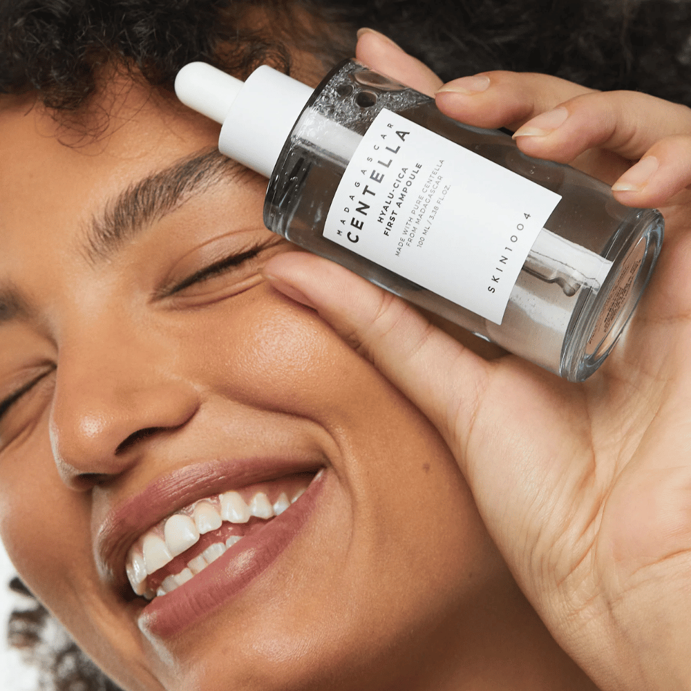 Hymyilevä nainen pitää Skin1004 Madagascar Centella Hyalu-Cica First Ampoule kasvojen vieressä.