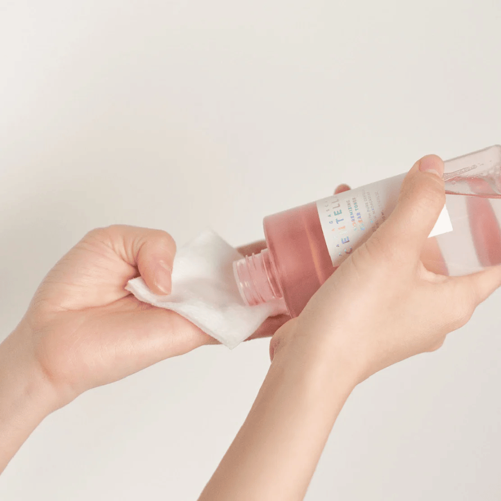 Henkilö kaataa Madagascar Centella Poremizing Clear Toner läpinäkyvästä pullosta vanulapulle. Tuotetta, joka on vaaleanpunainen, käytetään ihon virkistämiseen ja puhdistamiseen. Kädet ovat näkyvissä, mikä osoittaa tonerin levittämisen.