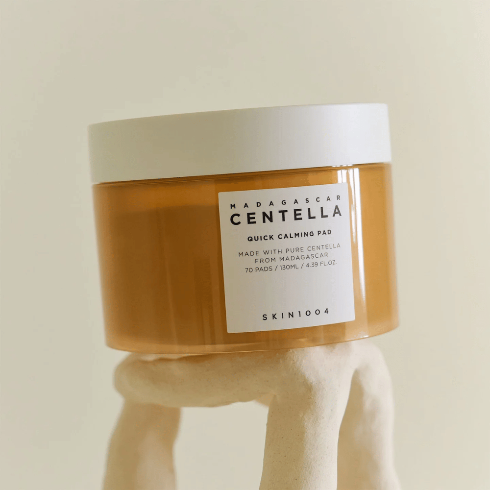 Purkki SKIN1004 Madagascar Centella Quick Calming Pad sijoitettu veistoksellisen valkoisen jalustan päälle – meripihkanvärinen säiliö, jossa on valkoinen etiketti ja kansi.