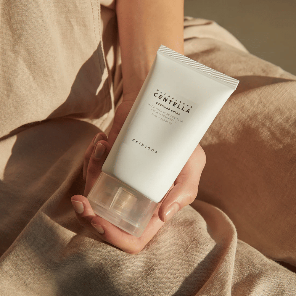 Henkilö pitää kädessään valkoista tuubia Madagascar Centella Soothing Cream SKIN1004. Tuubissa on läpinäkyvä korkki ja teksti, joka kuvaa tuotetta rauhoittavaksi ja valmistetuksi puhtaasta Centellasta. Henkilö on pukeutunut vaaleisiin vaatteisiin.