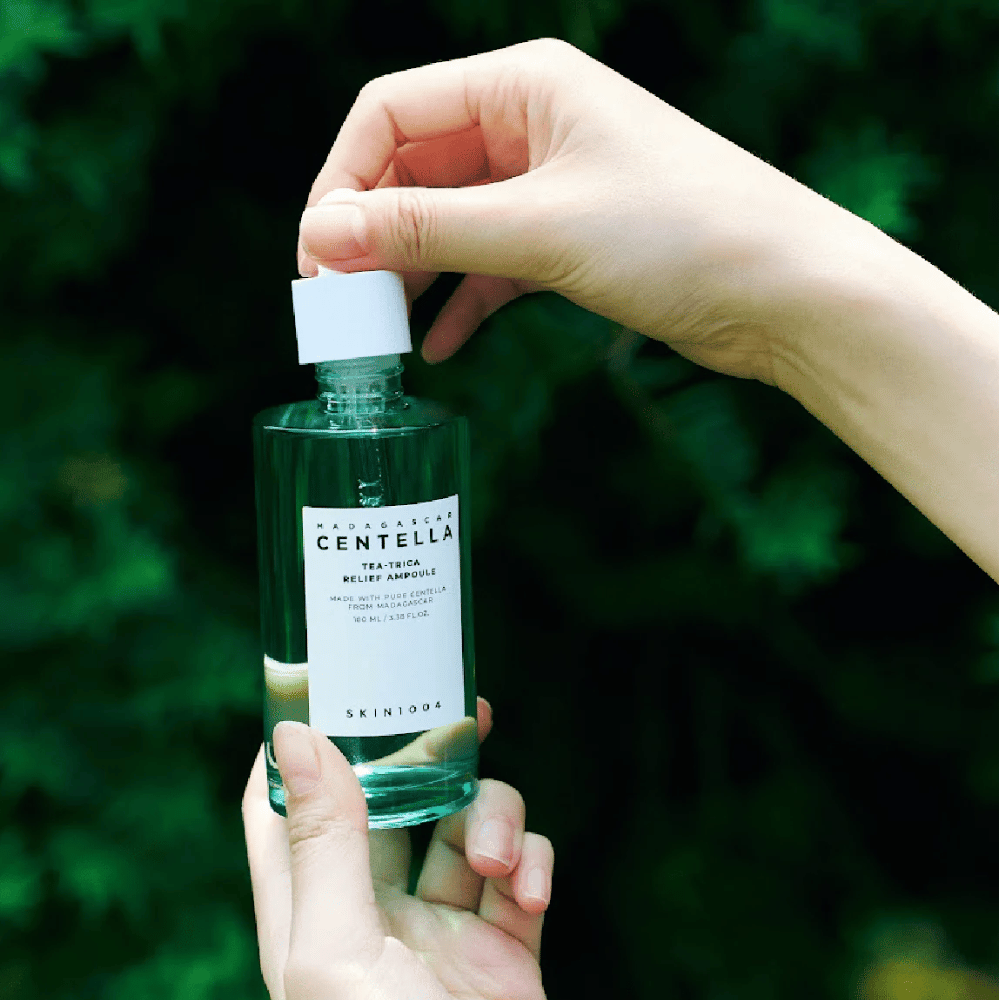Henkilö avaa SKIN1004 Madagascar Centella Tea-Trica Relief Ampoule – vihreä lasipullo, jossa on valkoinen etiketti, pidetään käsissä vihreää taustaa vasten.