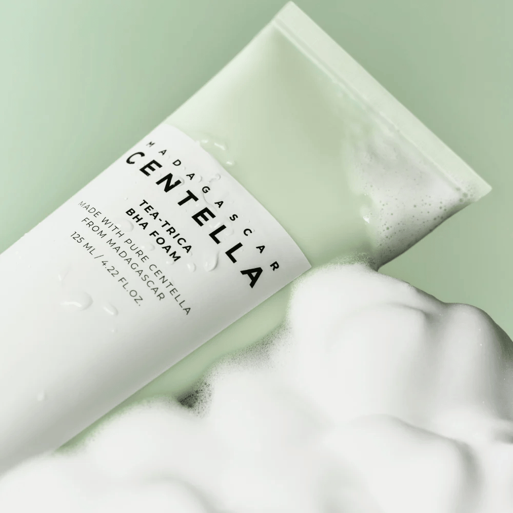 Puhdistusvaahtotyyppinen tuote, "Madagascar Centella Tea-trica BHA Foam," on osittain näkyvissä vaahdon ympäröimänä. Tuotteella on vihreä ja valkoinen muotoilu, ja tekstit "Madagascar Centella" ja "Tea-trica BHA Foam" ovat selvästi näkyvissä. Tilavuus on 125 ml.