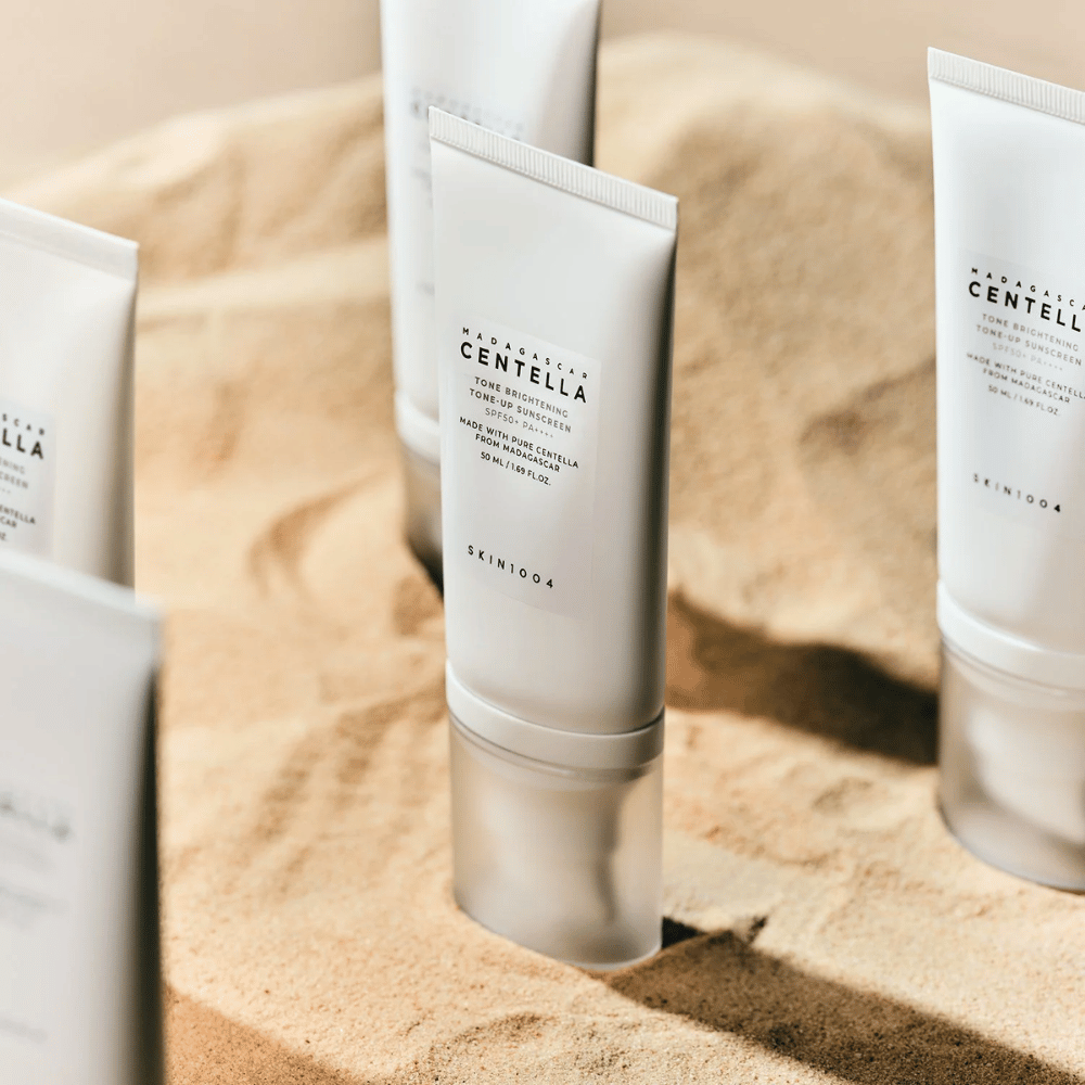 Useita putkiloita Madagascar Centella Tone Brightening Tone-Up Sunscreen seisoo hiekassa. Putkilo on valkoinen mustalla tekstillä ja sisältää 50 ml. Tuotteella on SPF 50+ PA++++ ja se on valmistettu puhtaasta Centellasta Madagaskarilta.