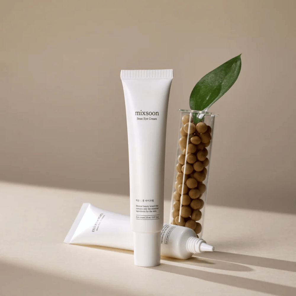 Kaksi Mixsoon Bean Eye Cream -tuubia sijoitettuna soijapavuilla täytetyn koeputken ja vihreän kasvinlehden viereen, beigellä pinnalla varjojen leikkiessä.
