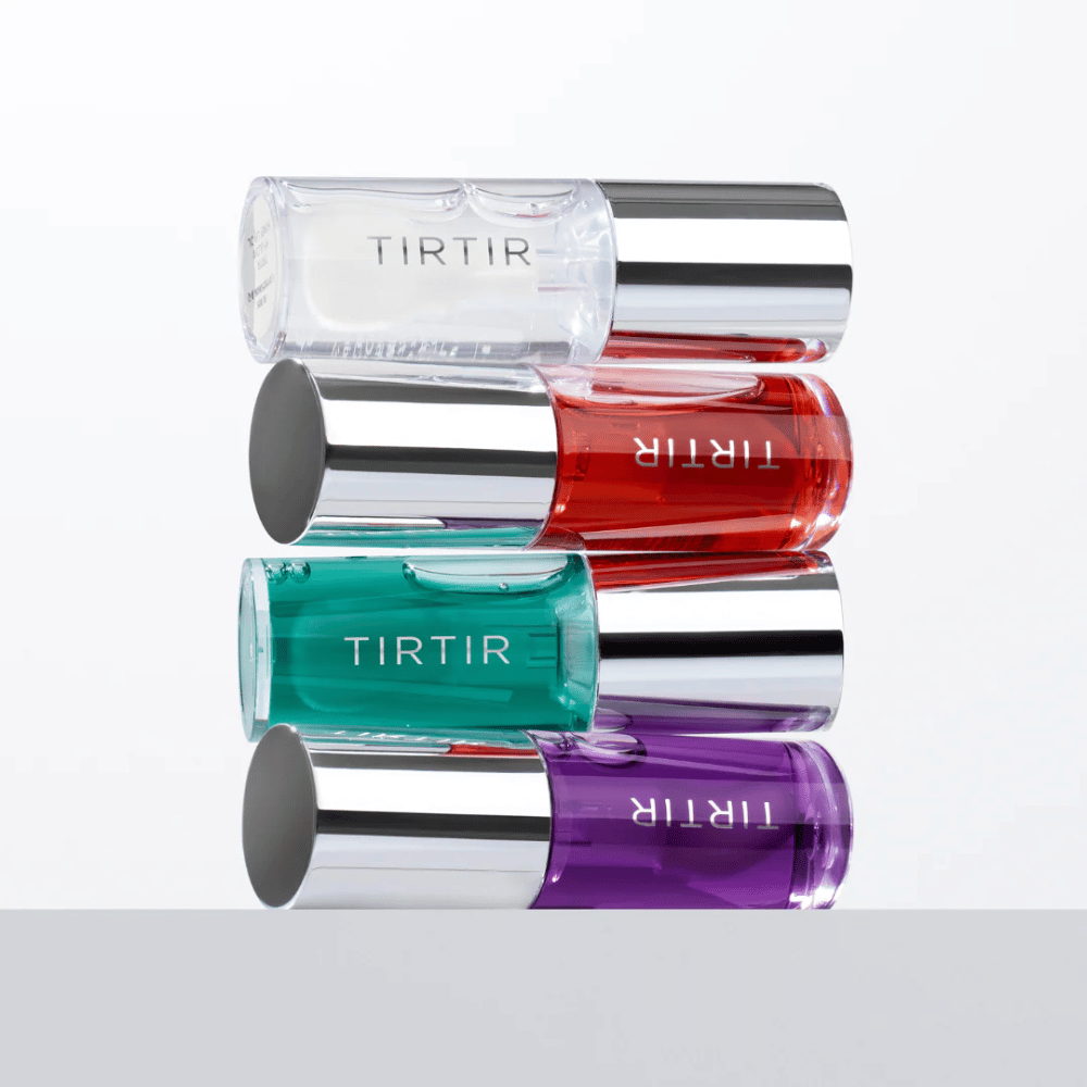 Neljä TIRTIR My Glow Lip Oil eri väreissä – läpinäkyvä, punainen, vihreä ja violetti – pinottuina toistensa päälle hopeanvärisillä kansilla valkoista taustaa vasten.